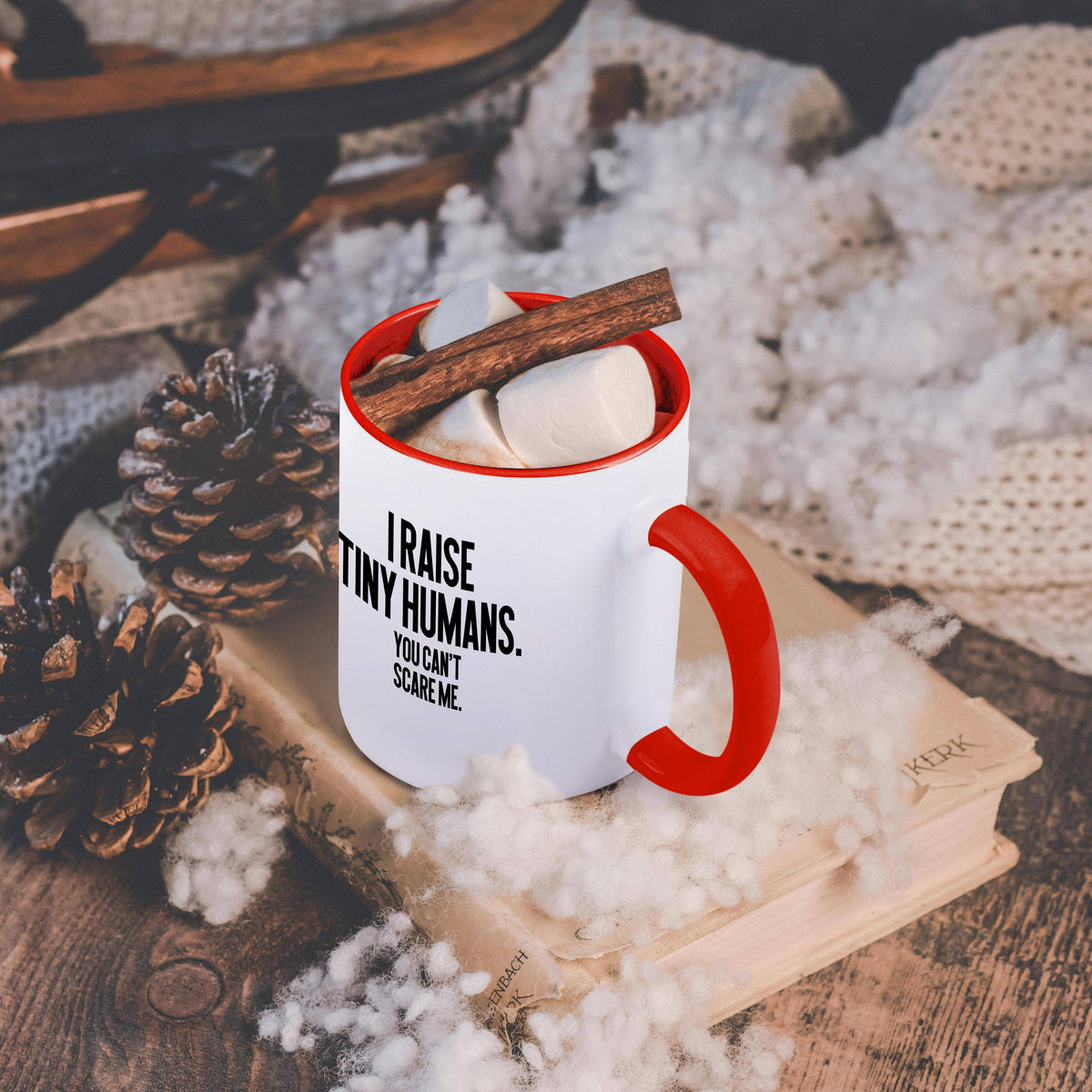 Red and white I Raise Tiny Humans funny mug, on a table with a cozy blanket