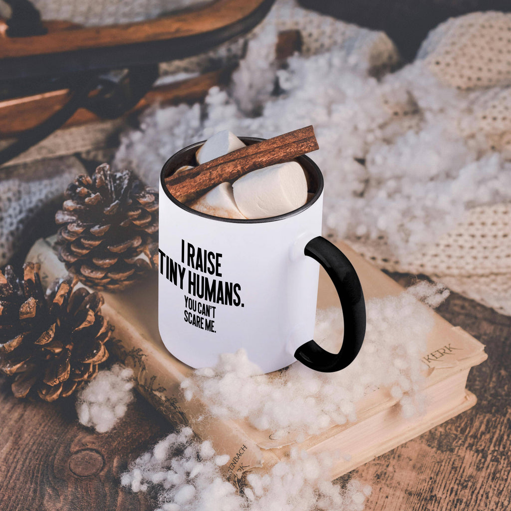 Black and white I Raise Tiny Humans 15 oz mug, on a table with a cozy blanket