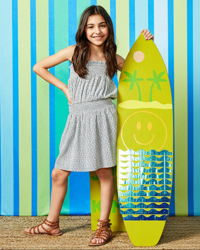 Tween girl in a light sage green sun dress holding a surfboard, perfect for sunny days shopping, at school, or to the beach.